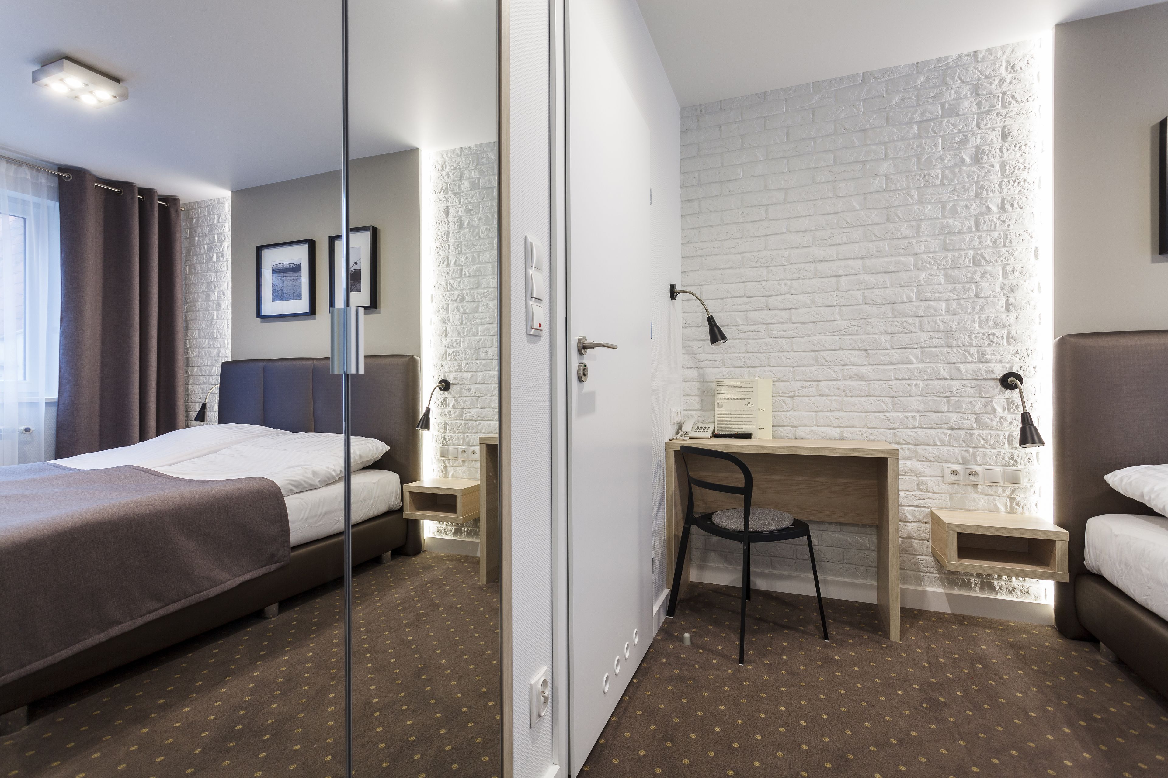 Habitación de hotel con cama, escritorio y silla. Se observa una pared de ladrillo blanco y una ventana con cortinas.