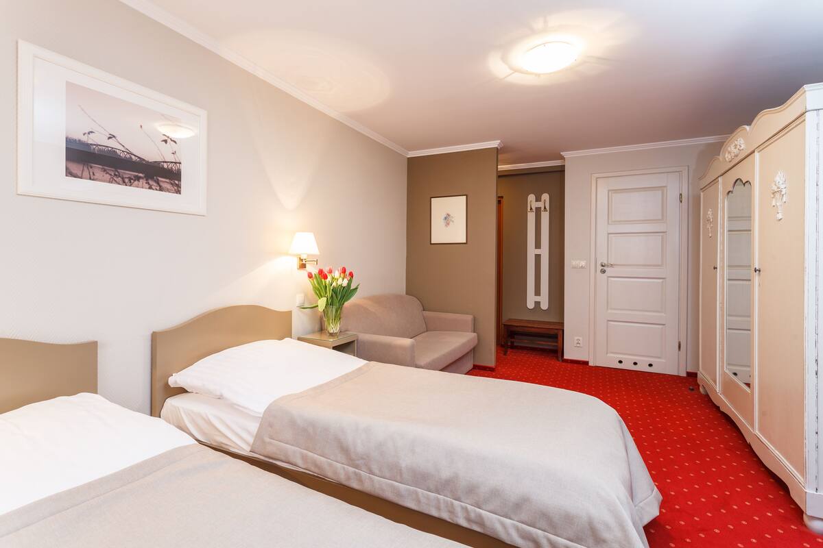 A hotel room with two beds, a red carpet, a desk with a lamp, a chair, and a wardrobe.