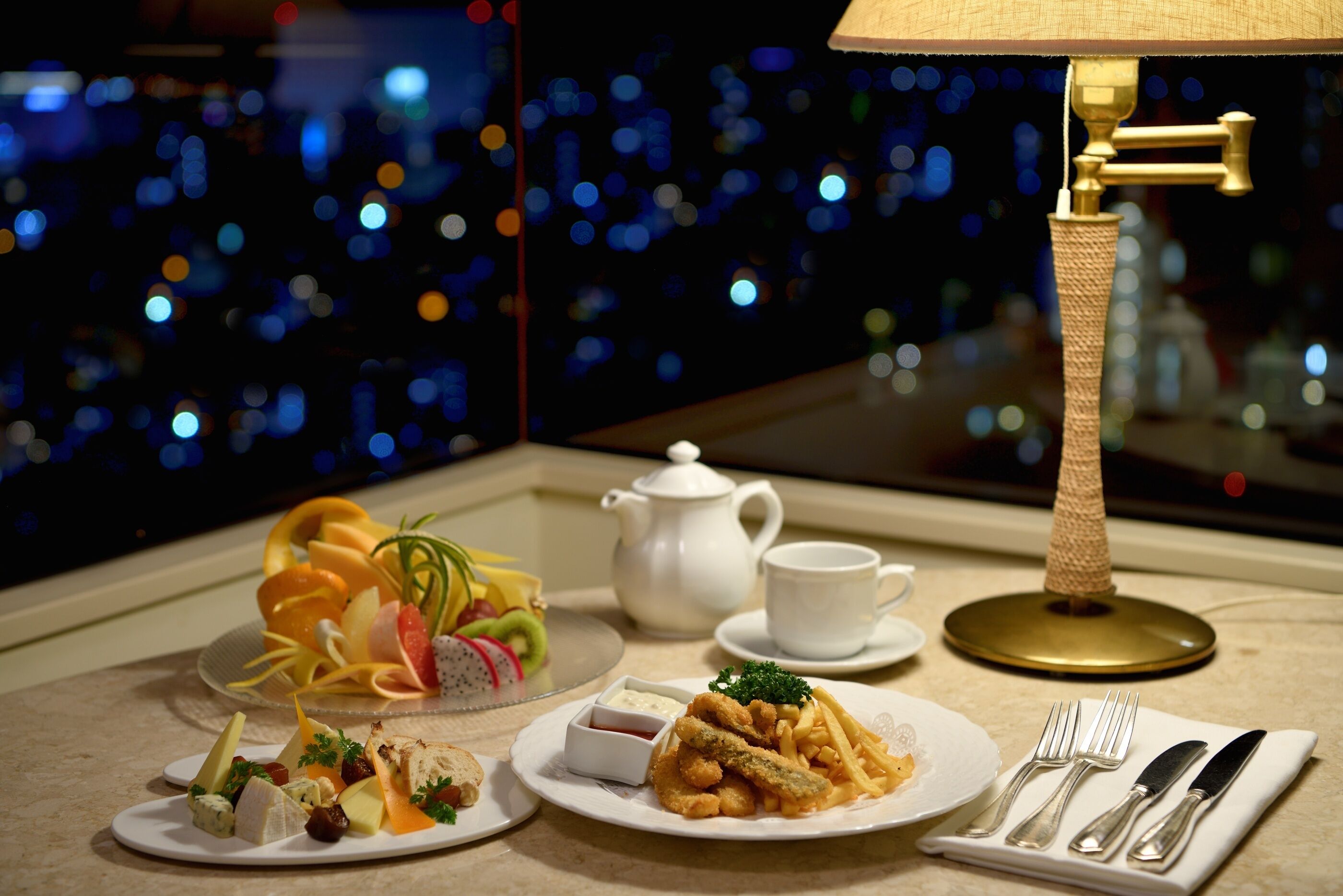 Ein Esstisch, gedeckt mit Speisen, einer Teekanne, einer Tasse und einer Lampe, mit Stadtblick im Hintergrund.