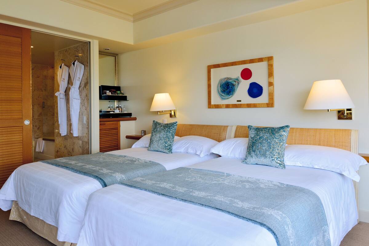 A hotel room with two beds, a wooden door, a bathroom visible through an open door, and a wall-mounted artwork.