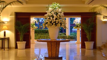 Um hall com um grande arranjo de flores brancas sobre um pedestal, duas plantas em vasos e uma luminária sobre uma mesa.
