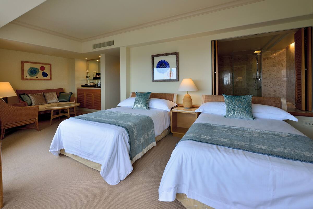 A hotel room with two beds, a sofa, a small table, and a bathroom with a glass shower.