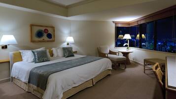 A hotel room with a large bed, two chairs, a small table, and a window with a city view.