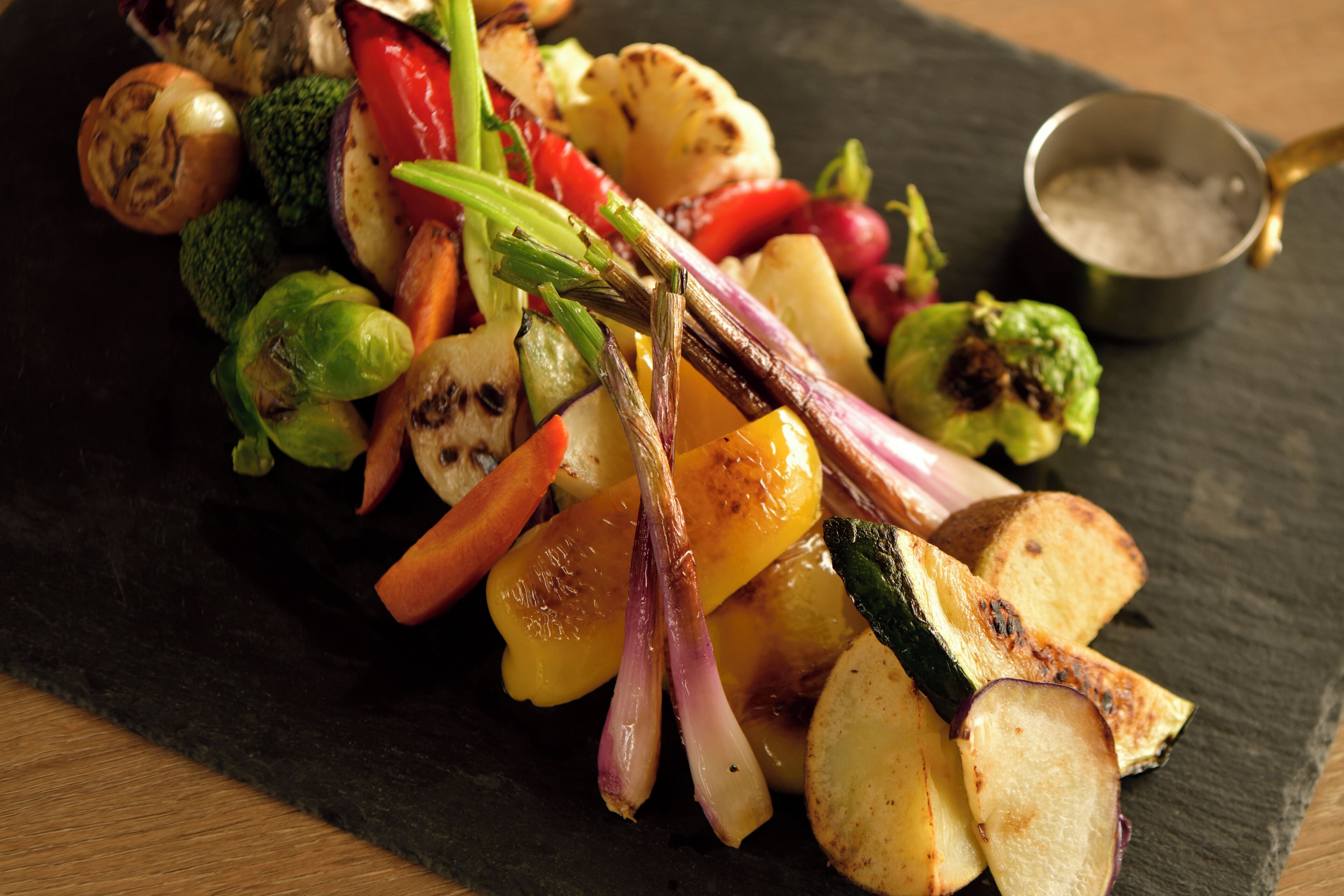 A variety of grilled vegetables and a small bowl of salt on a slate serving board.