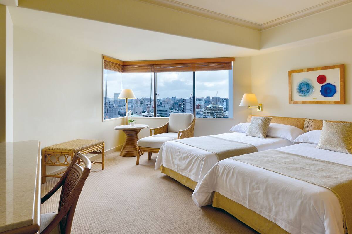 A hotel room with two beds, a desk, a chair, a window with a city view, and a wall art piece.