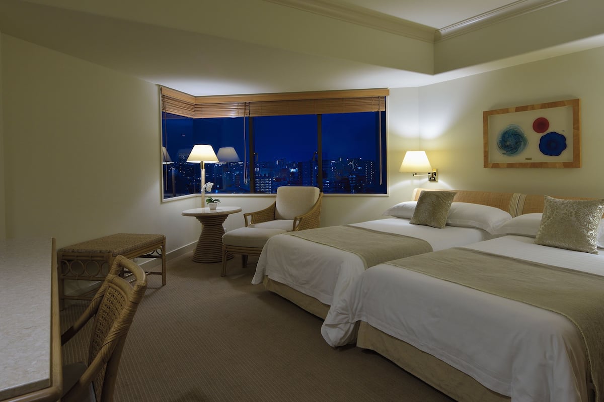 A hotel room with two beds, a sitting area with a table and chairs, and a large window with a city view.