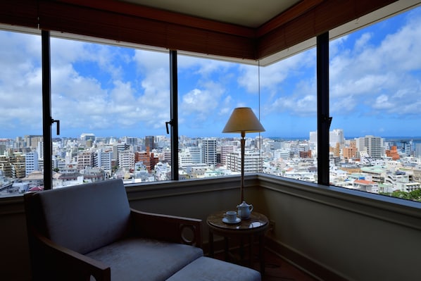 A room with a large window offering a city view, a chair, a small table with a lamp, and a cup. - The Naha Terrace (Naha)