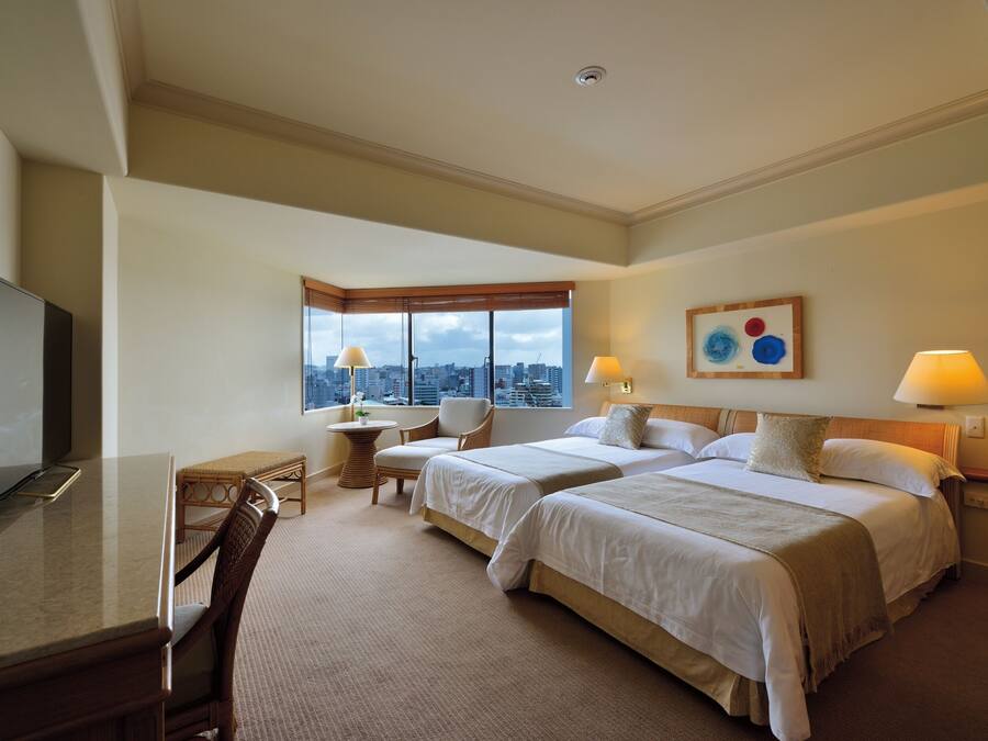 A hotel room with two beds, a desk, a chair, a television, and a window with a city view.