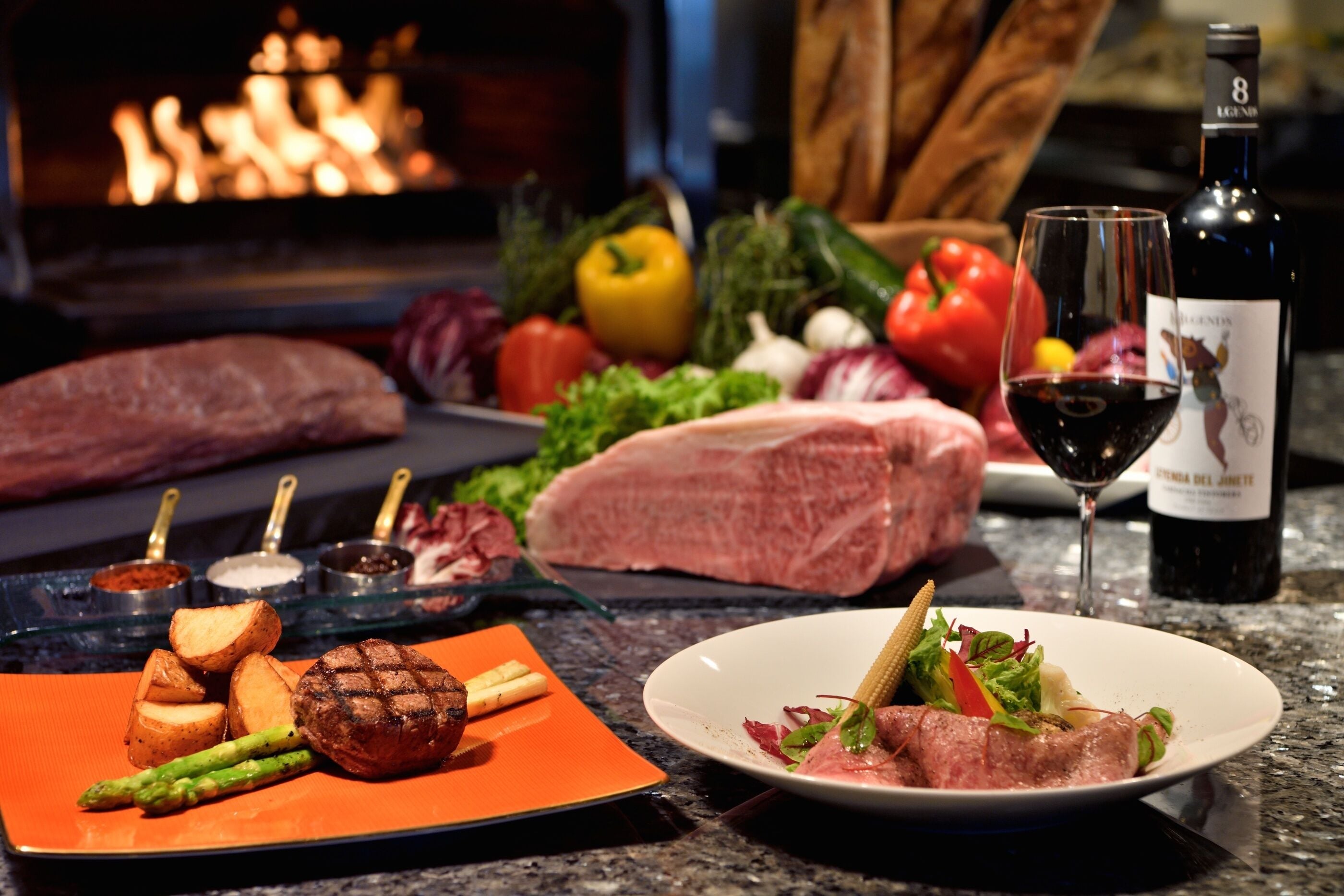 A dining table with various meats, vegetables, and a bottle of wine.
