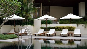 Zona de piscina con sillones de descanso y sombrillas, rodeada de vegetación y un edificio moderno.
