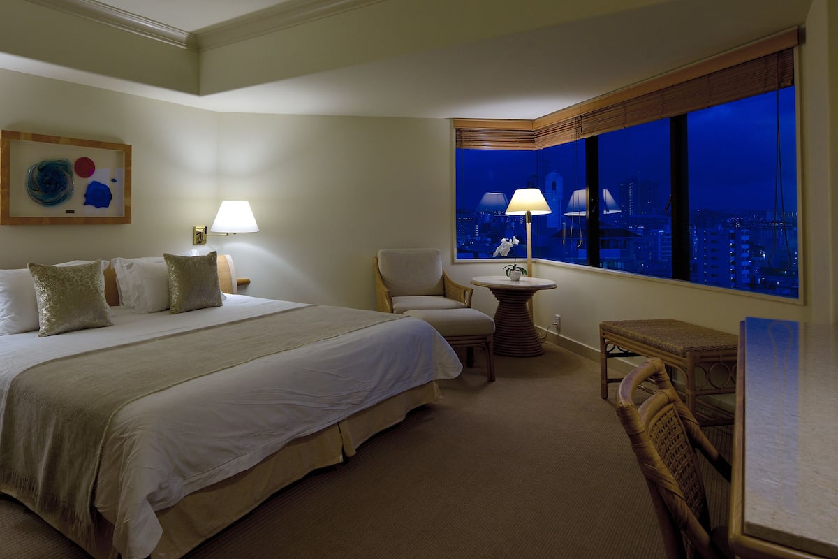 A hotel room with a large bed, two chairs, a small table, and a view of the city at night.