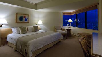 Habitación de hotel con una cama grande, dos sillas, una mesita y vista a la ciudad de noche.