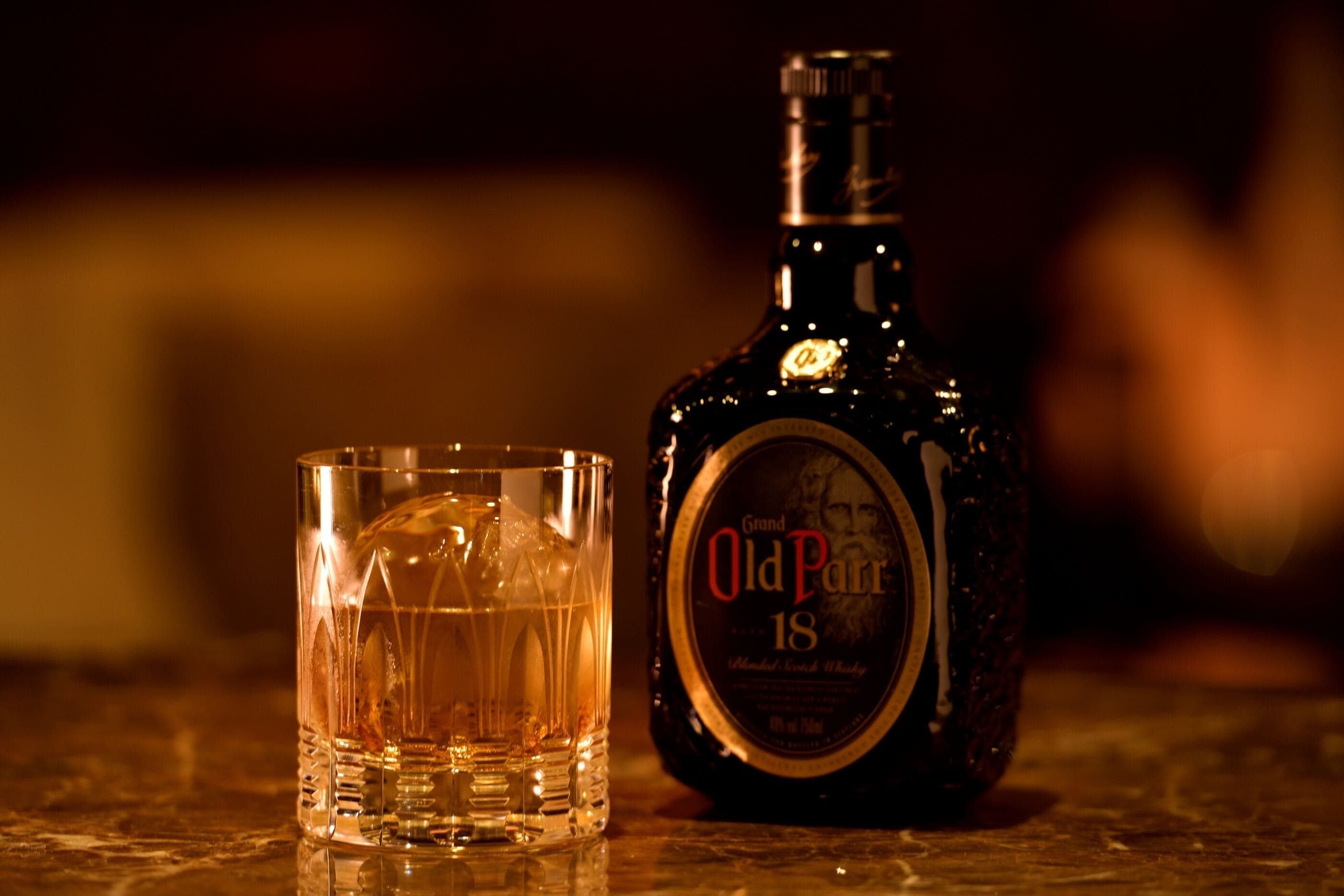 A bottle of Grand Old Parr 18 whisky and a glass on a reflective surface.