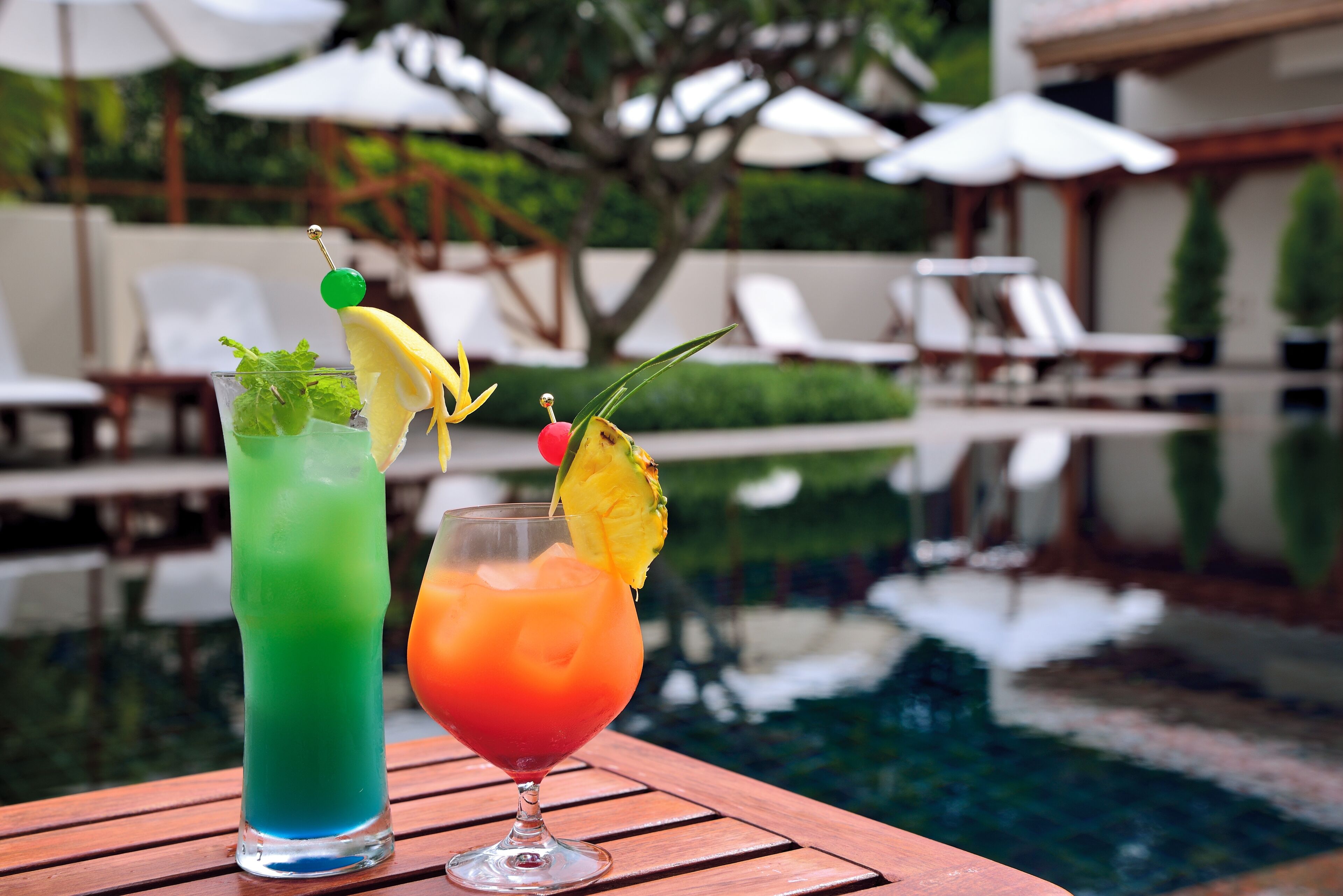 Two cocktails on a wooden table by a poolside with lounge chairs and umbrellas in the background.