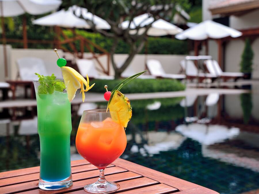 Two cocktails on a wooden table by a poolside with lounge chairs and umbrellas in the background.