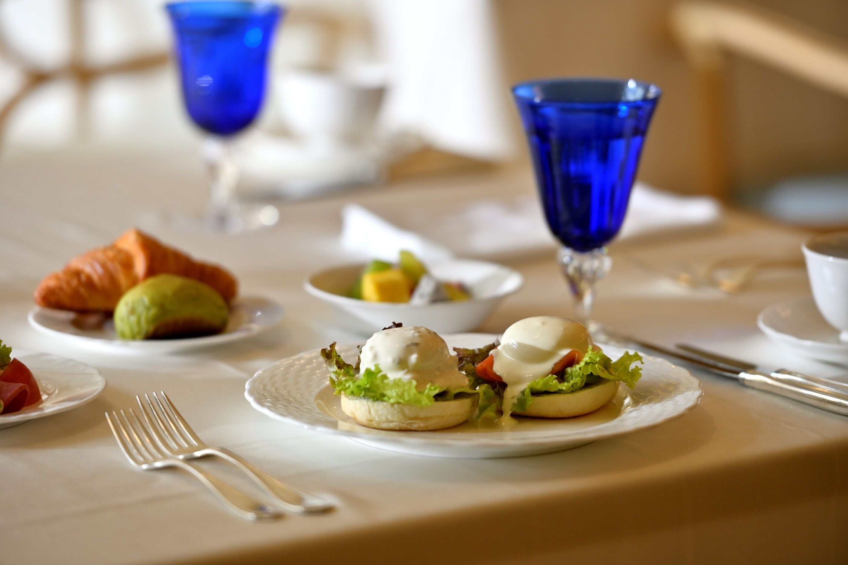 A table set with plates of food, including eggs benedict and a croissant, and blue glassware.