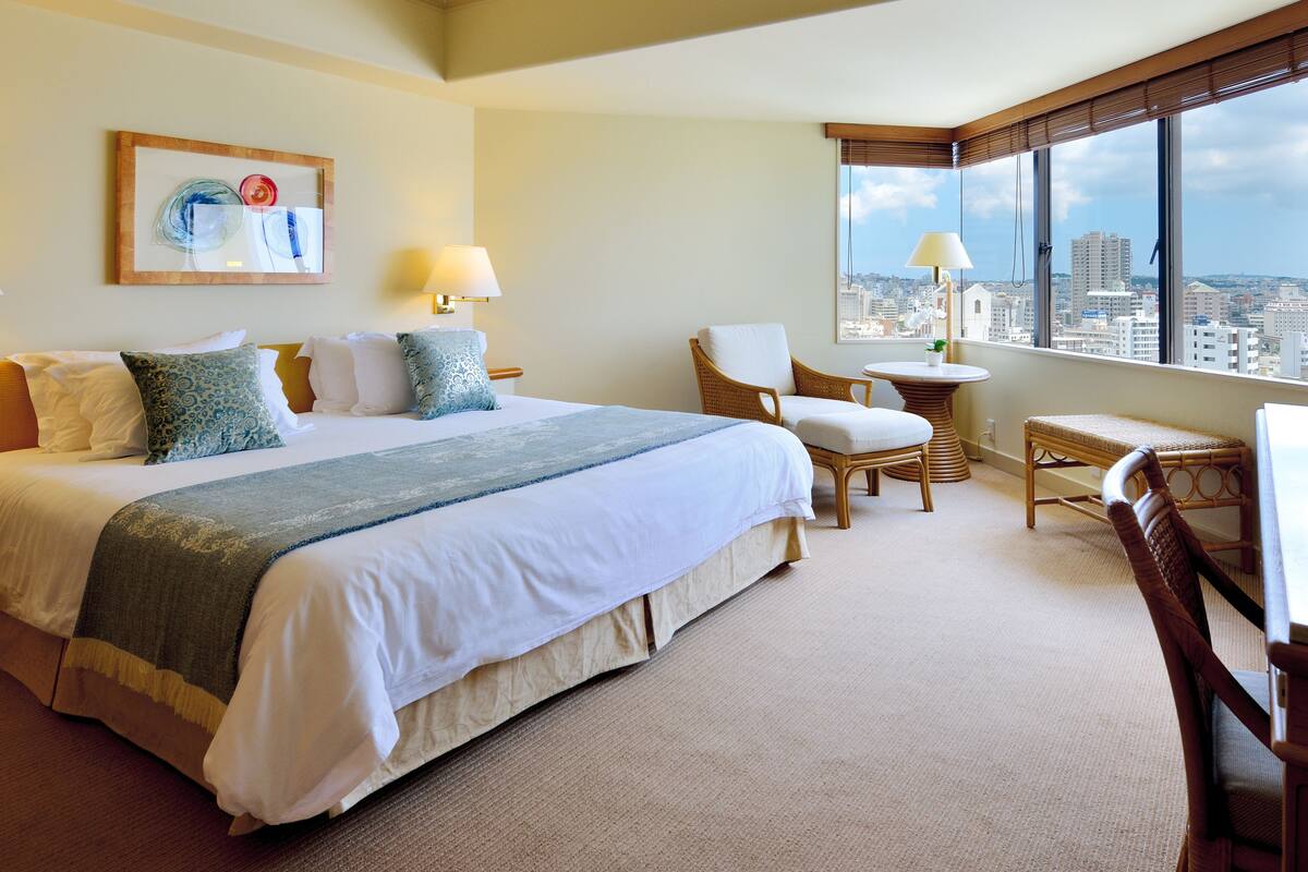 A hotel room with a large bed, two chairs, a small table, and a city view through the window.