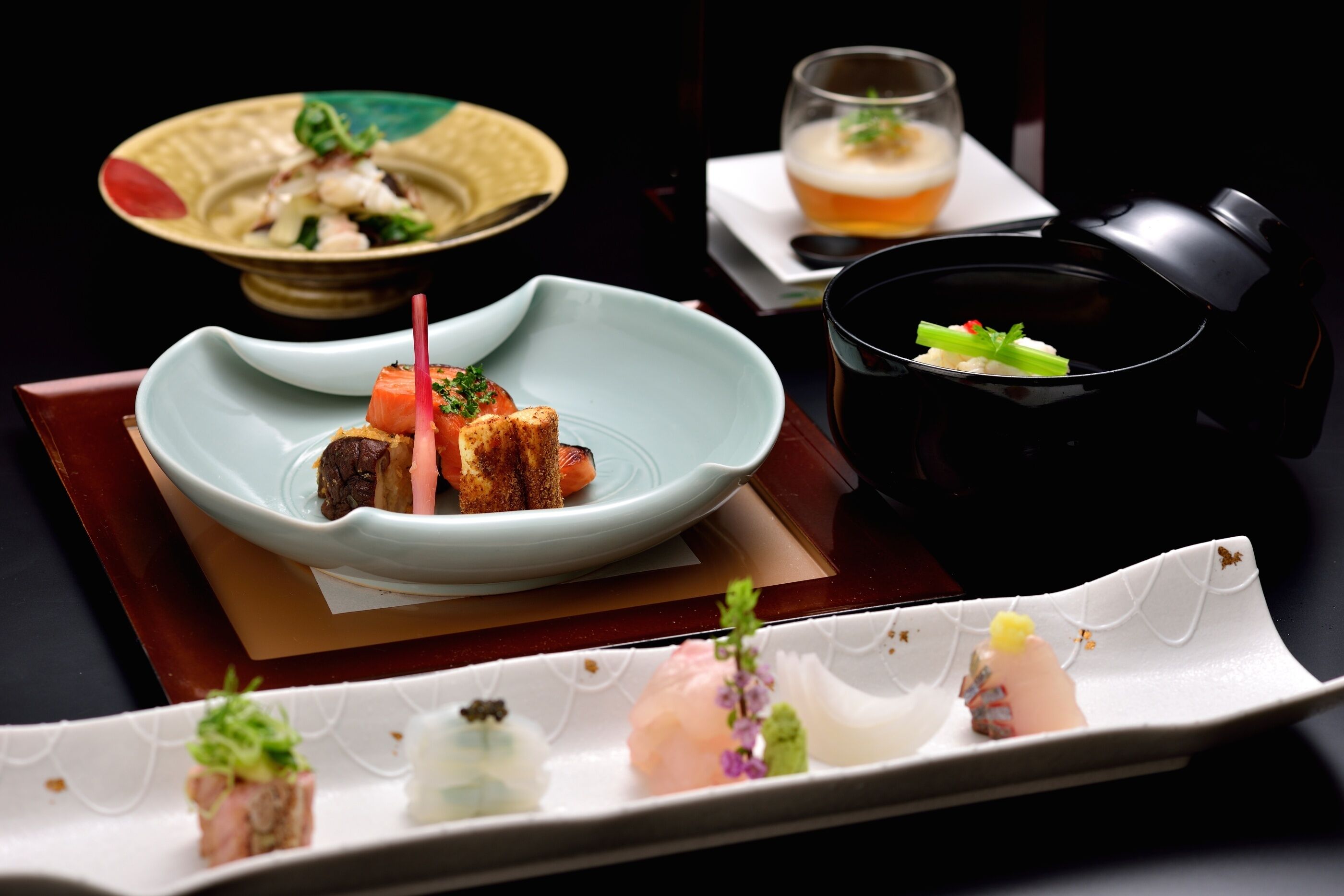 A variety of dishes on a table, including a bowl of food, a glass of beer, and a black bowl with a green garnish.