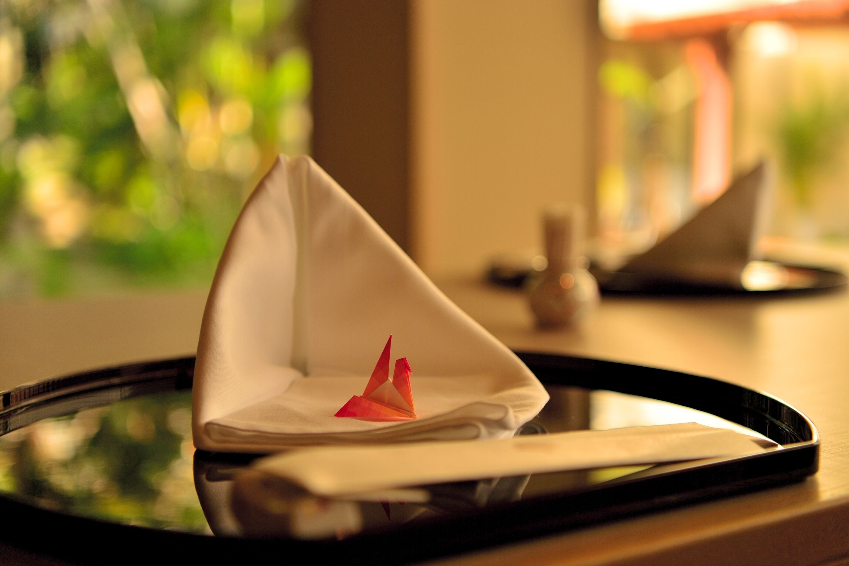 A tray with a folded napkin and a red origami crane on a reflective surface.