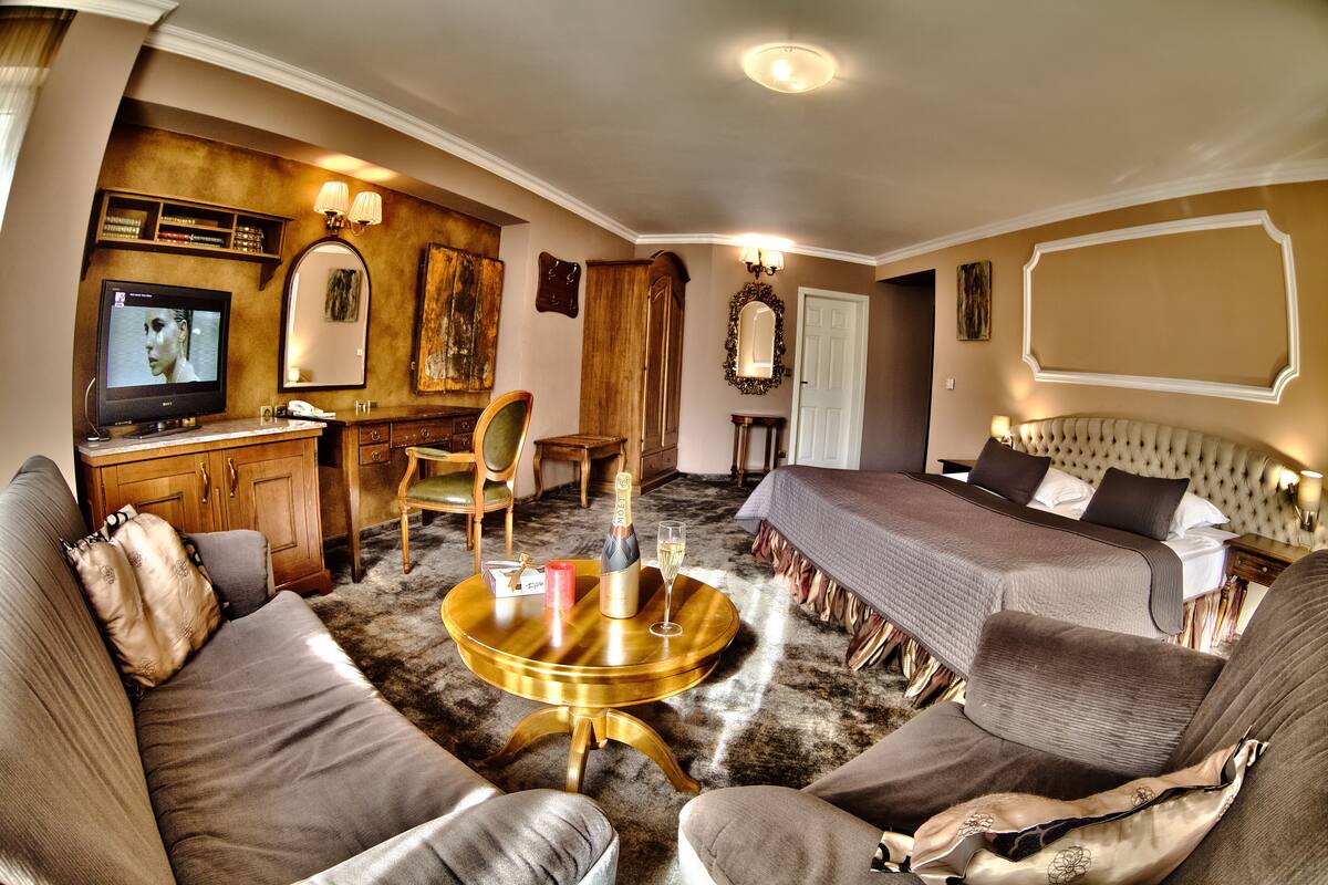 A hotel room with a sofa, two single beds, a small table with a bottle and glass, a TV, a wooden cabinet, and a mirror.