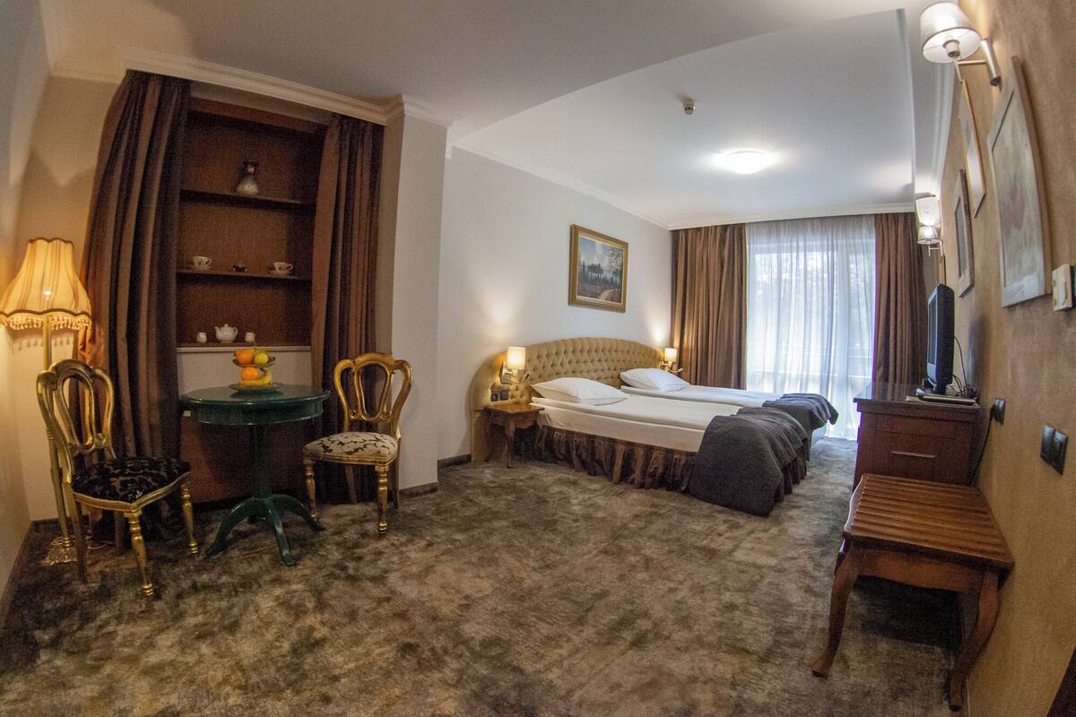 A hotel room with a large bed, two bedside tables with lamps, a wooden chair, a small table with a lamp, a shelf with decorative items, and a painting on the wall.