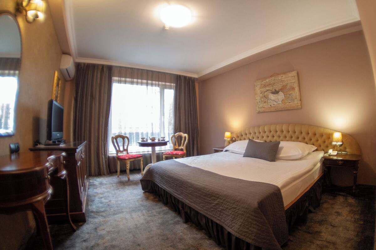 A hotel room with a large bed, a desk with a television, two chairs, a small table, and a window with curtains.