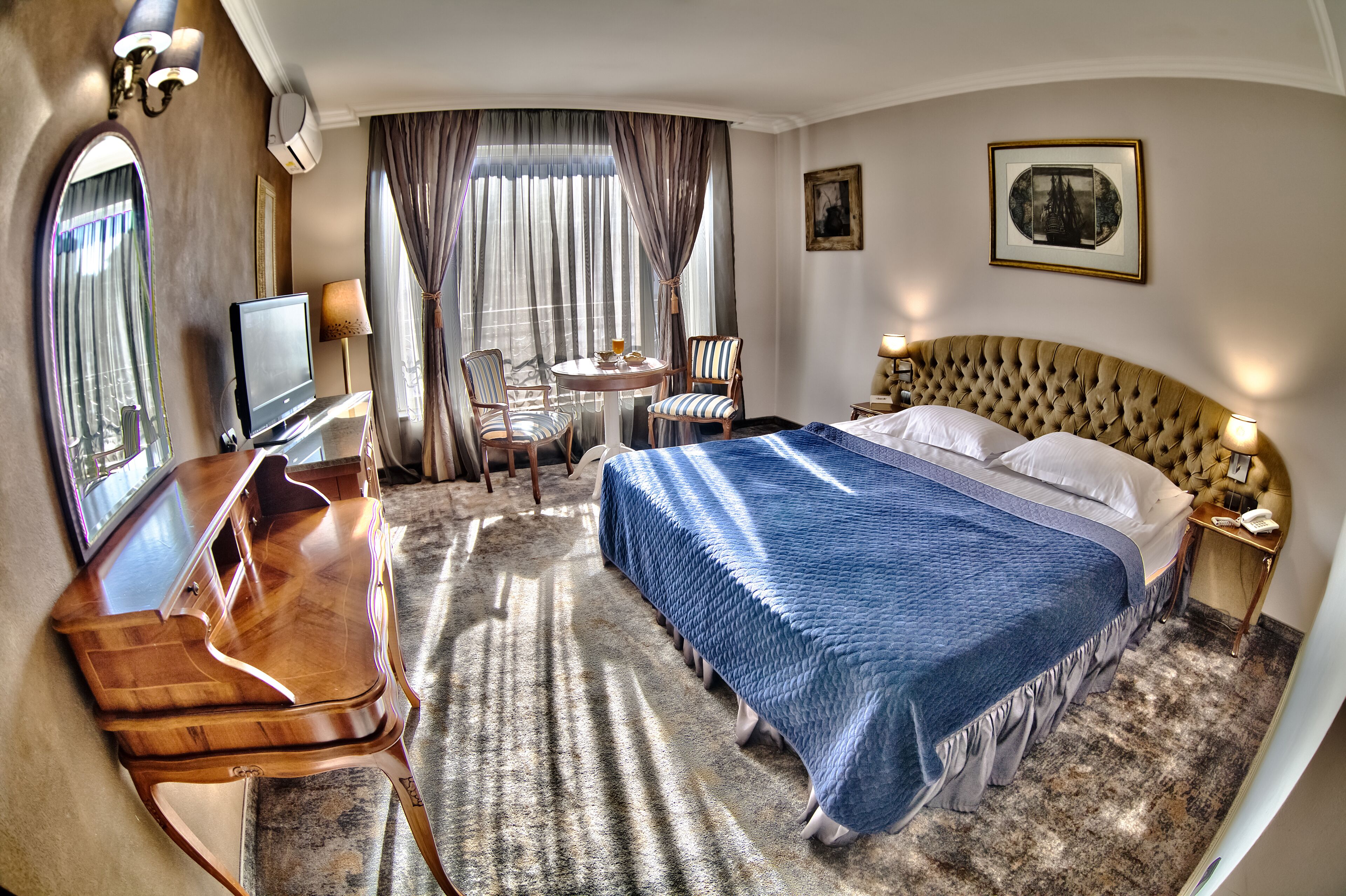 A hotel room with a large bed, a desk with a television, a small round table with chairs, and a window with curtains.
