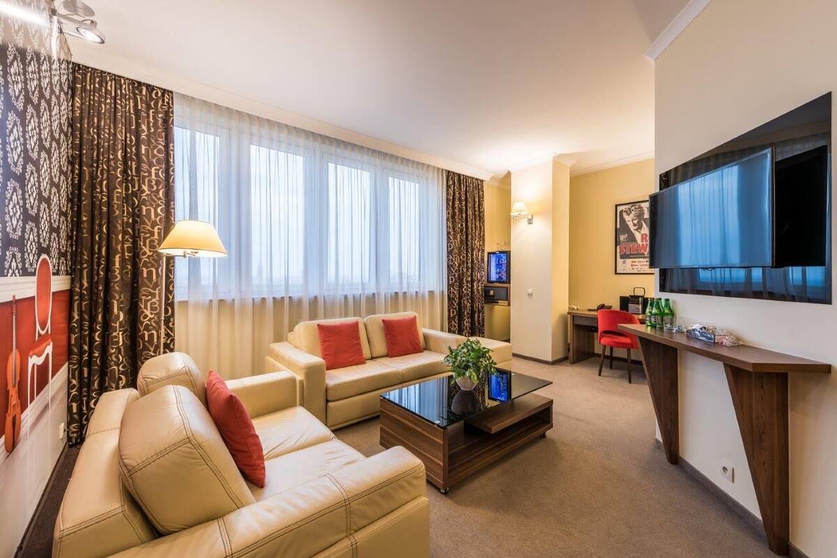 A hotel room with a living area featuring a sofa, a coffee table, and a TV. There is a dining area with a table and chairs, and a kitchen area with a microwave and a refrigerator.
