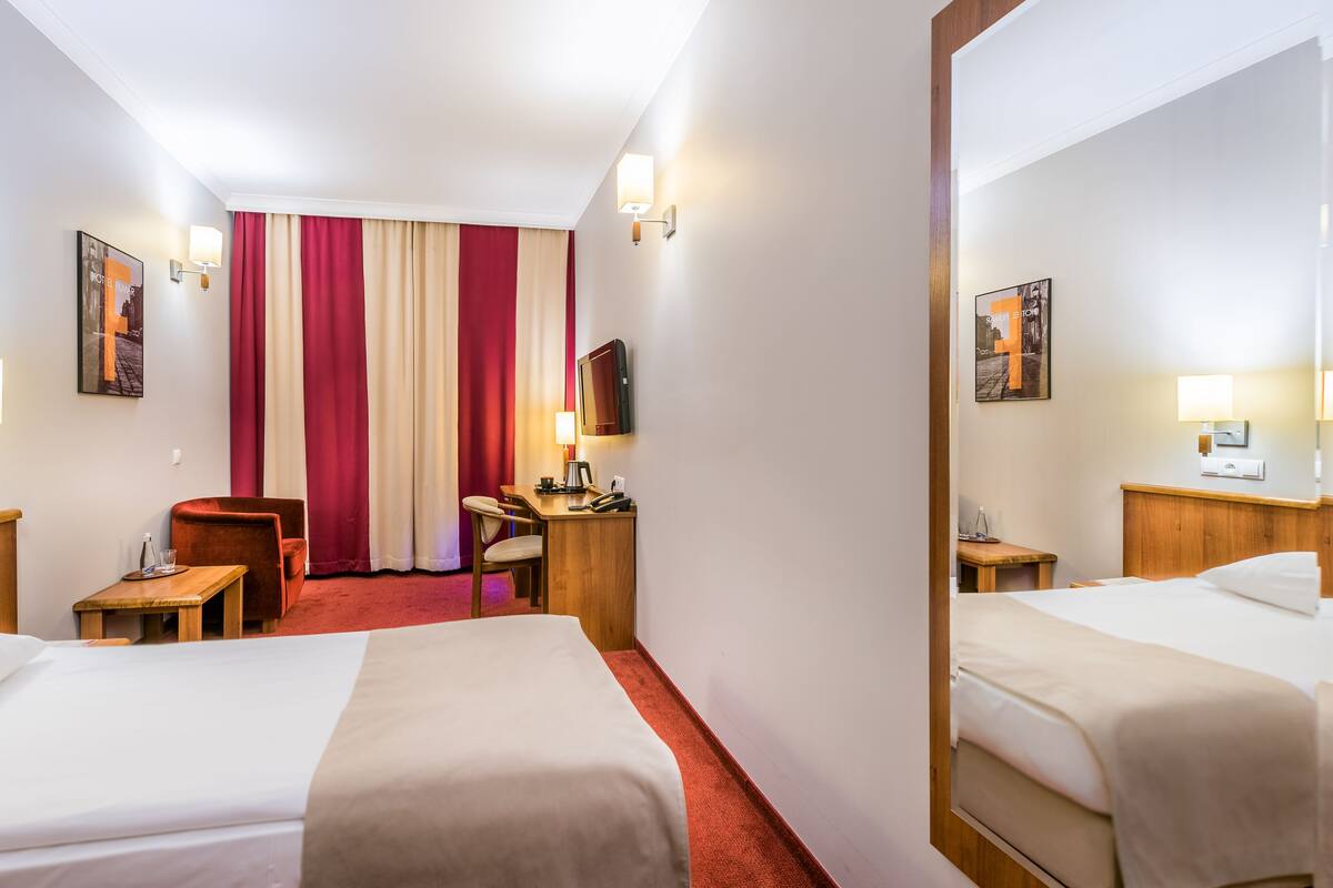 A hotel room with two beds, a desk with a chair, a TV, and a window with red and white curtains.