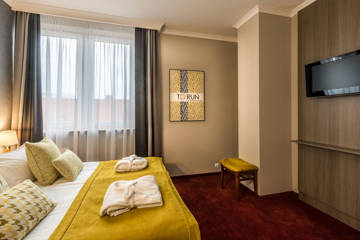 A hotel room with a bed, a TV mounted on the wall, a small stool, and a framed artwork.
