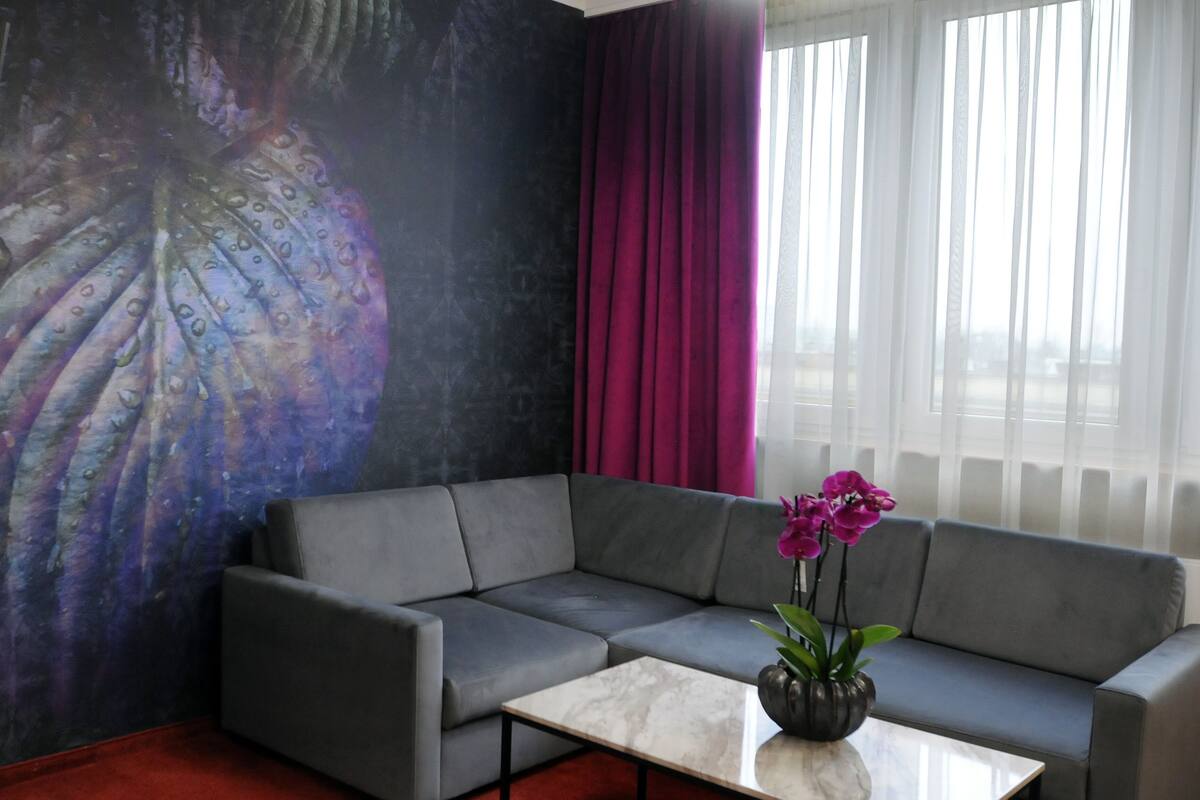 A modern living room with a grey sectional sofa, a marble coffee table, a large abstract painting on the wall, a purple orchid vase, and a window with sheer white curtains.