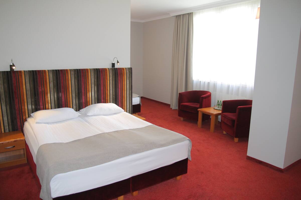 A hotel room with a large bed, a desk, two chairs, and a window with curtains.