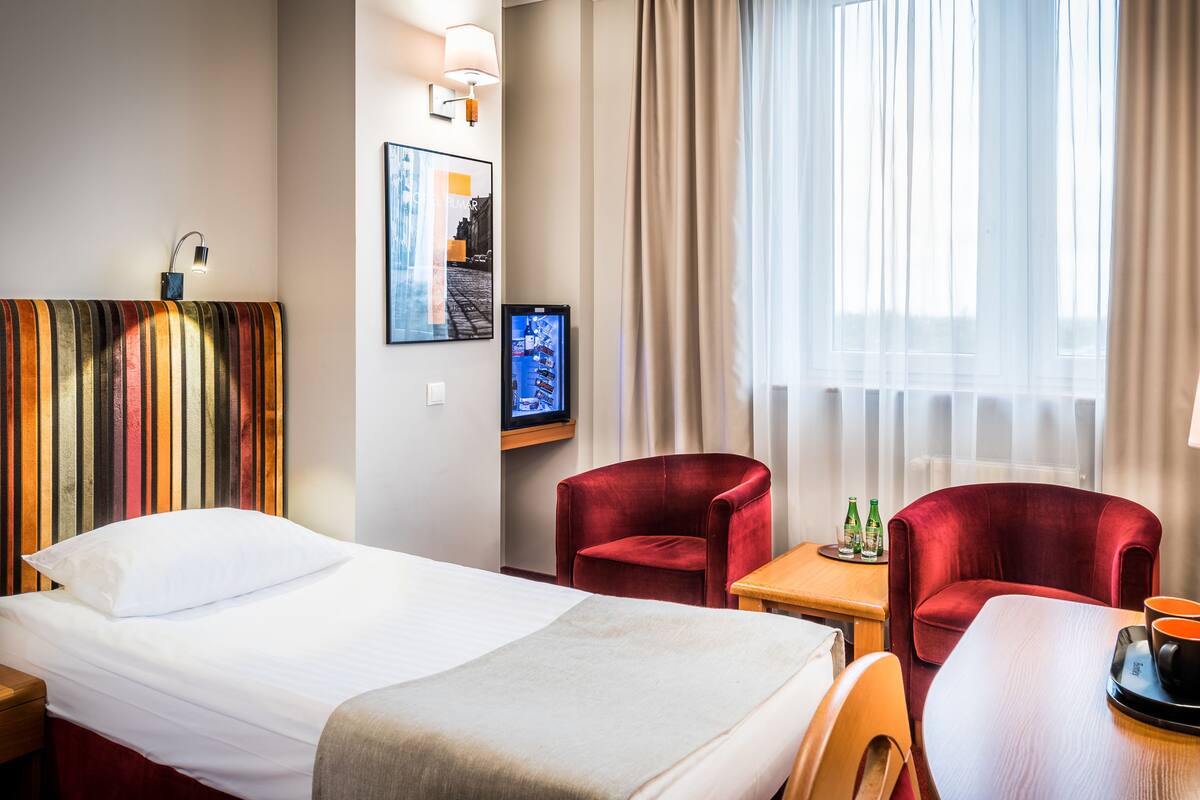 A hotel room with a bed, two red armchairs, a small table with a coffee maker and cups, and a TV on a stand.