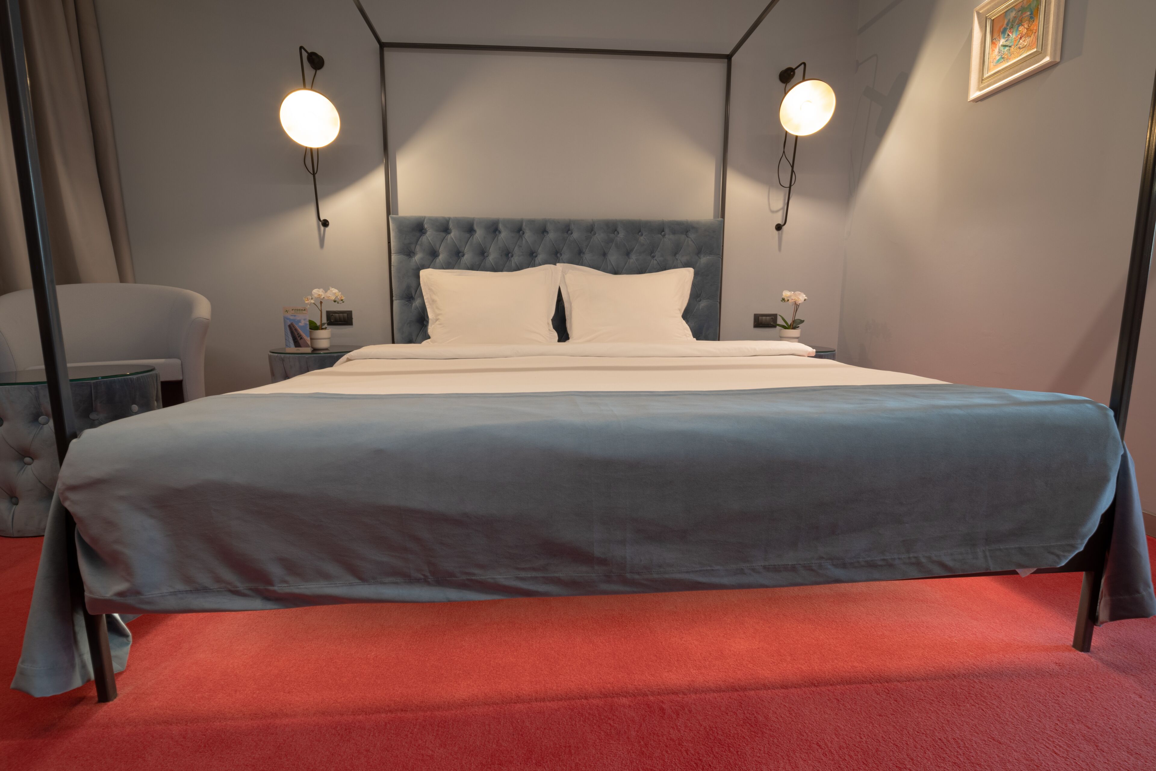 Una cama bien tendida con una colcha azul y blanca, dos mesitas de noche con lámparas, un sillón tapizado de gris y una alfombra roja.