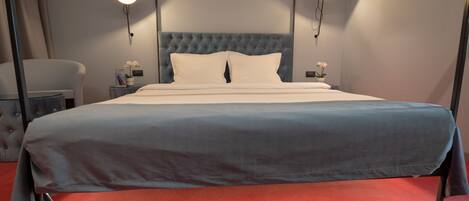 Una cama bien tendida con una colcha azul y blanca, dos mesitas de noche con lámparas, un sillón tapizado de gris y una alfombra roja.