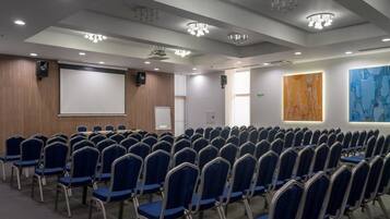 Een congreszaal met rijen blauwe stoelen, een projectiescherm en een groot abstract schilderij aan de muur.