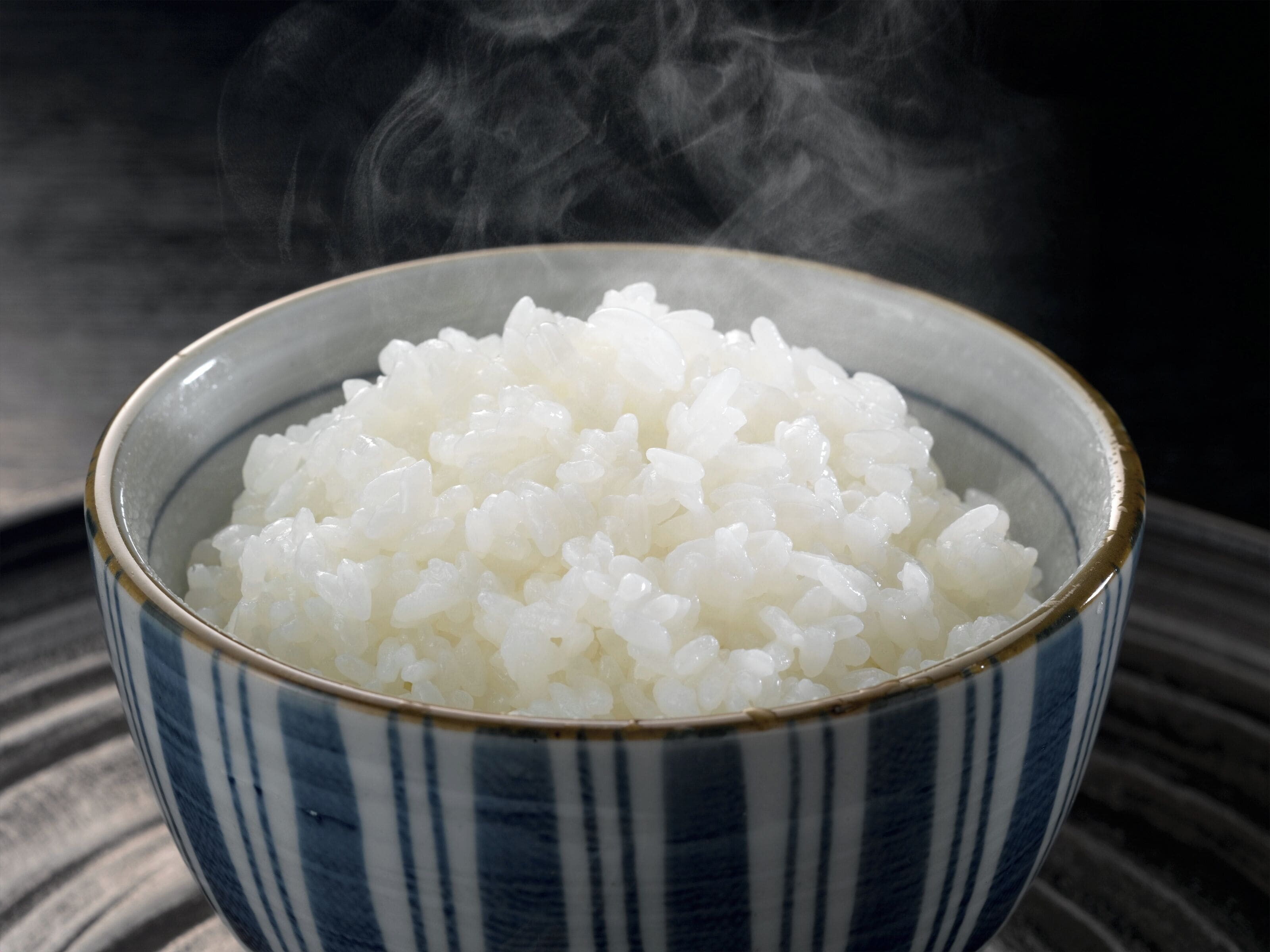 A bowl of white rice with steam rising from it, placed on a striped surface.