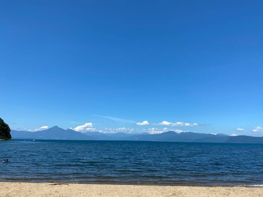 A beach with clear blue water, a sandy shore, and distant mountains under a clear sky.