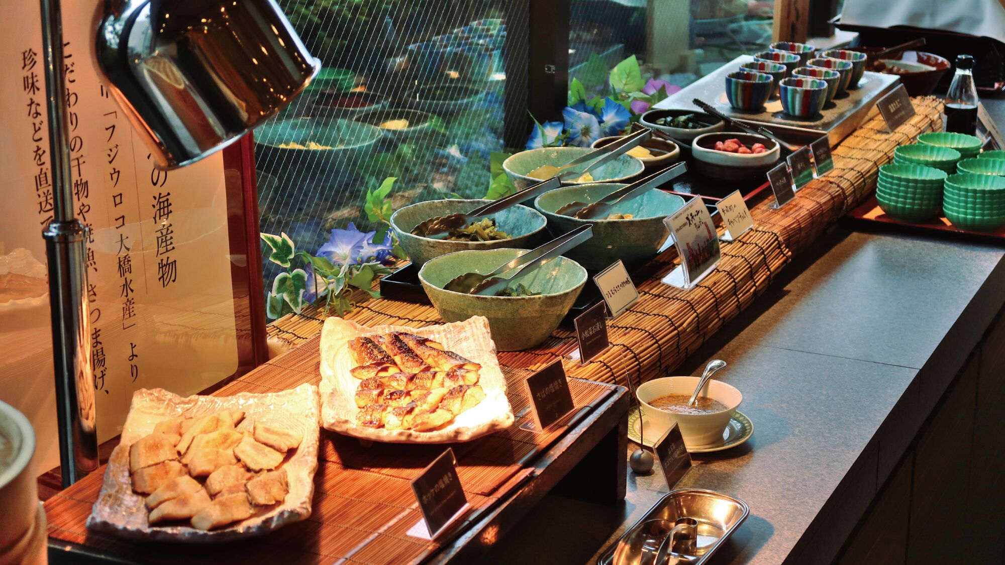 Ein Buffet mit verschiedenen Speisen und Schalen, ein Schild mit japanischem Text und eine Zimmerpflanze im Hintergrund.