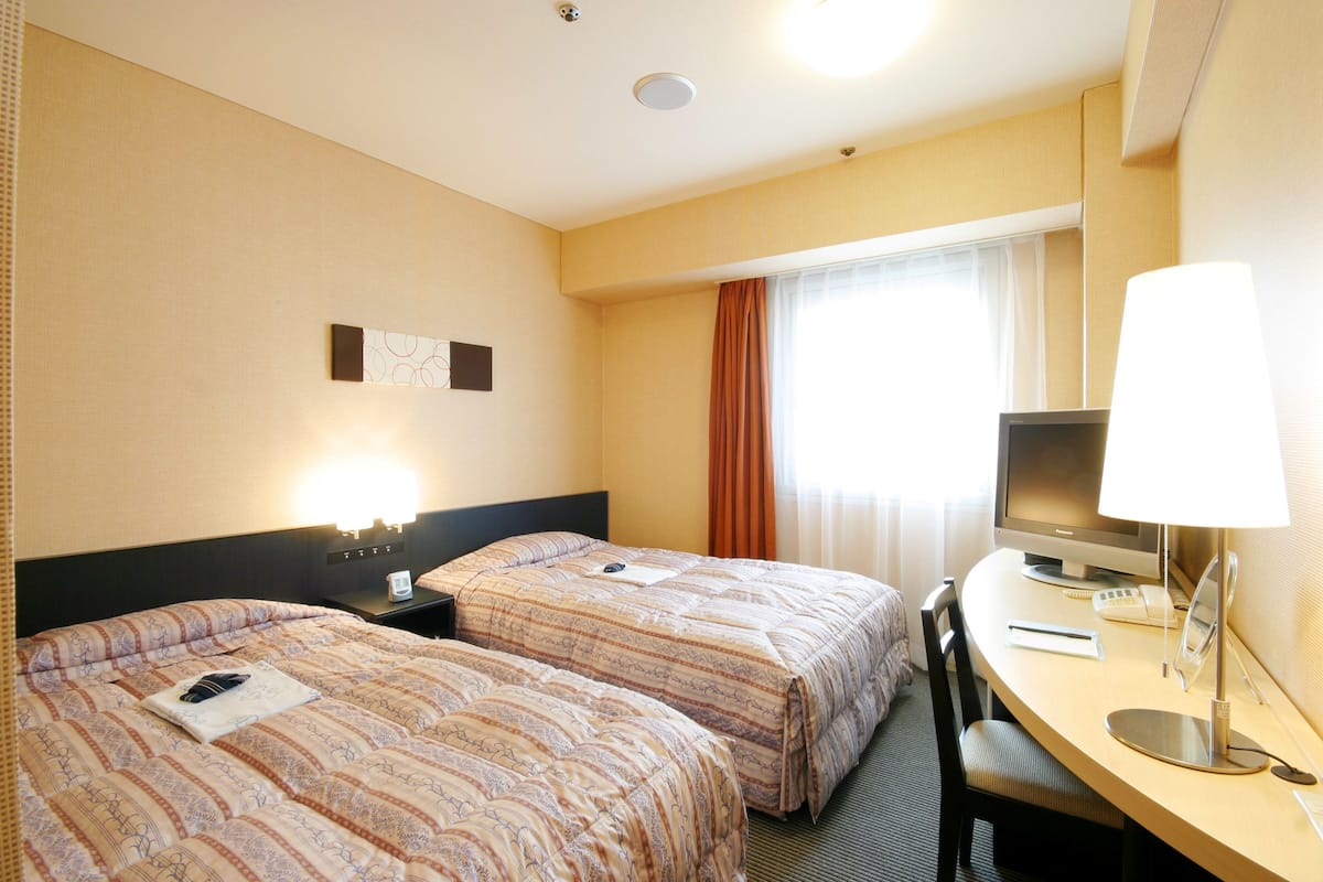 Une chambre d’hôtel avec deux lits, un bureau avec un ordinateur et une fenêtre avec des rideaux.