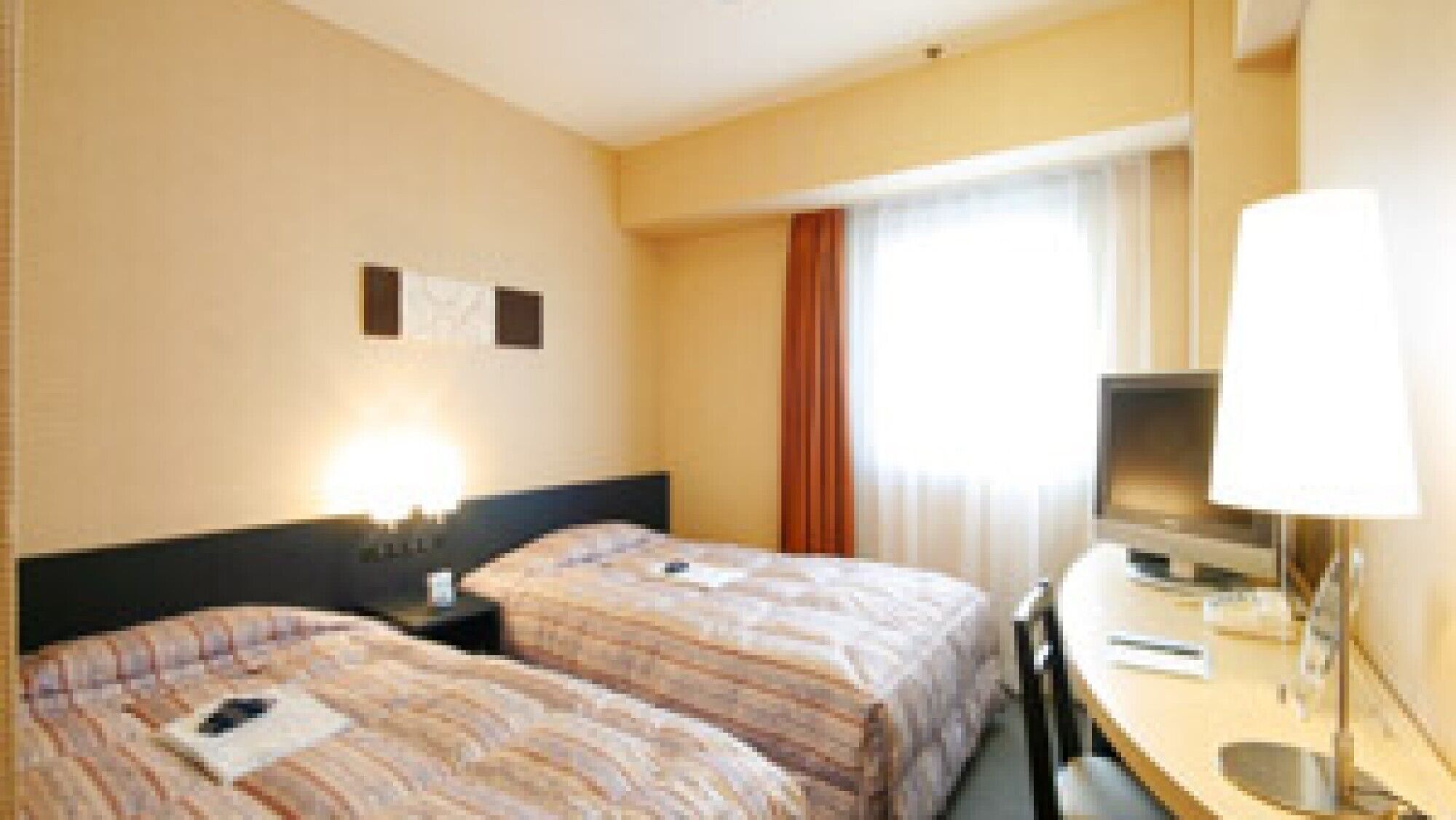 Et hotelværelse med to senge, et skrivebord med stol, et fjernsyn og et vindue med gardiner.