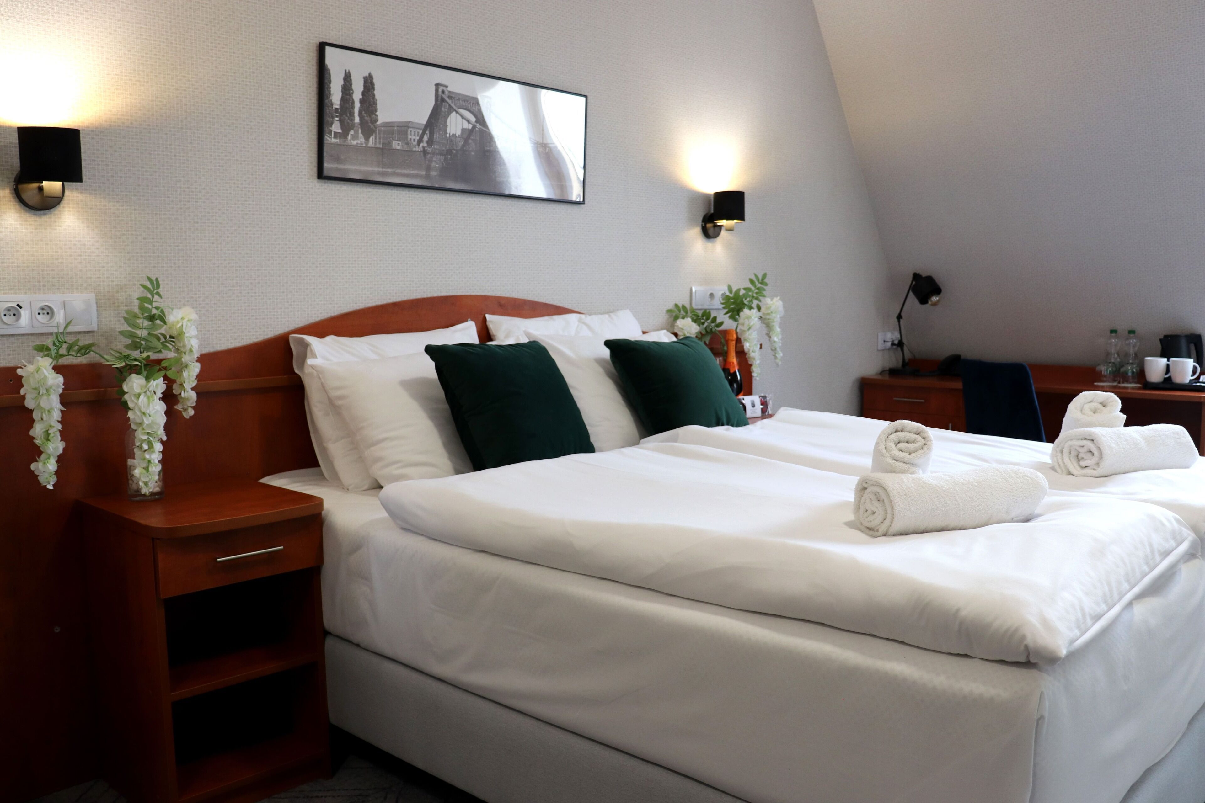 Quarto de hotel com uma cama grande, duas mesas de cabeceira, um criado-mudo, um quadro na parede e duas arandelas.