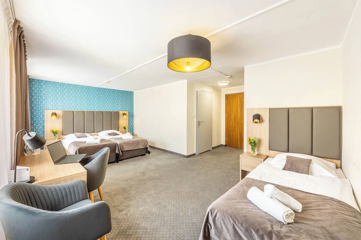 A hotel room with two beds, a desk with a laptop, a chair, a lamp, and a large window.