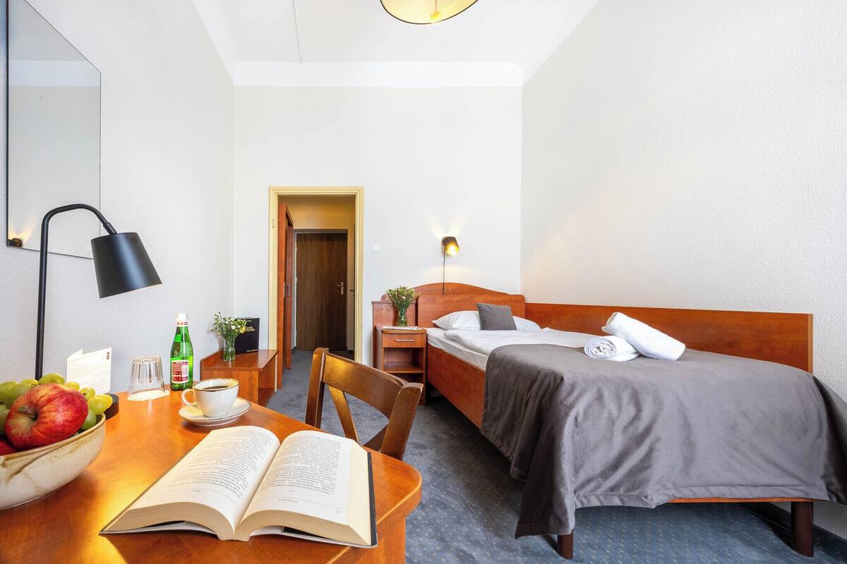 A hotel room with a wooden bed, a bedside table with a lamp, a bowl of fruit, and an open book on a wooden desk.