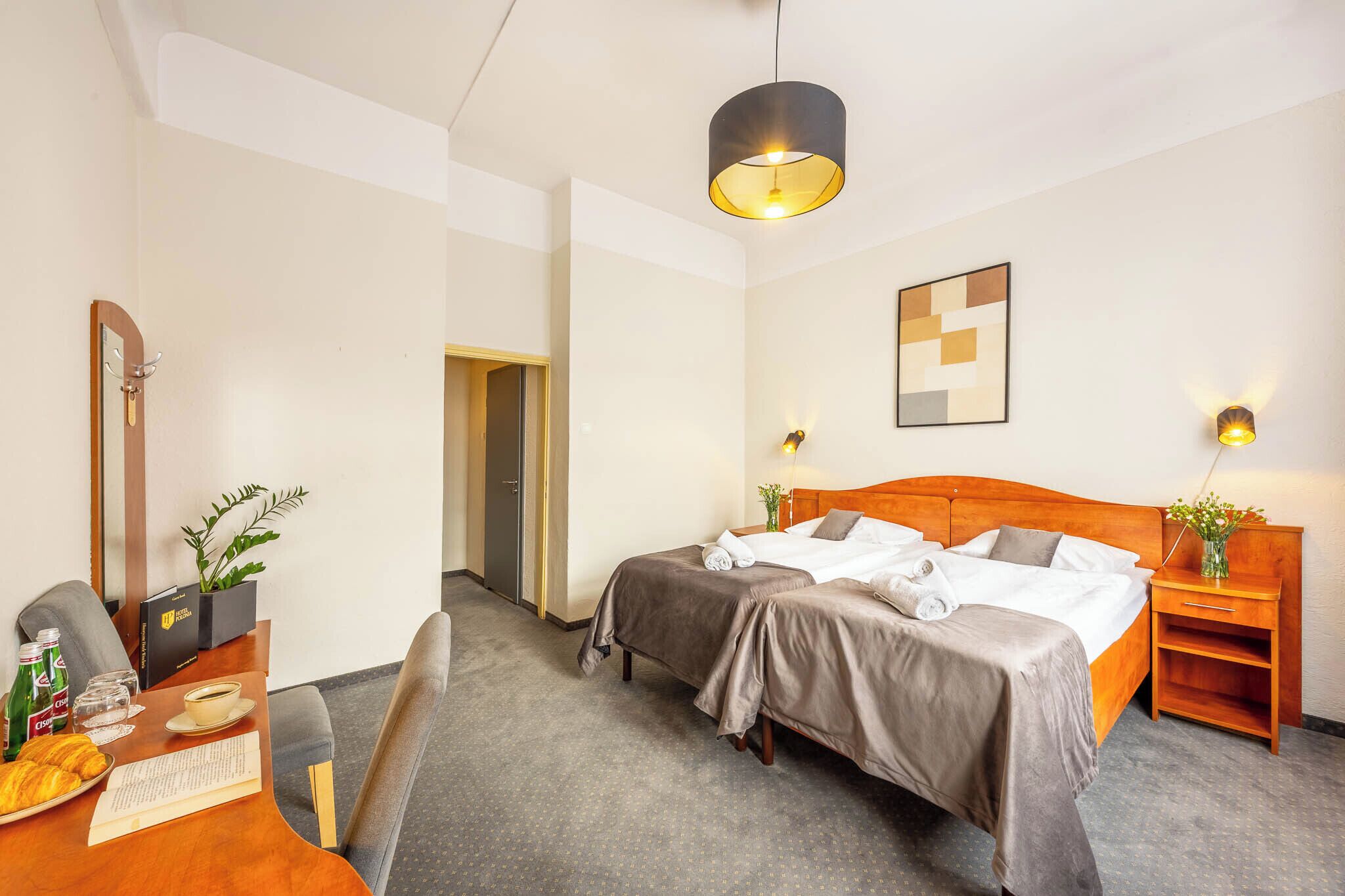 A hotel room with a double bed, a desk with a chair, a coffee table, a plant, and a wall-mounted light fixture.
