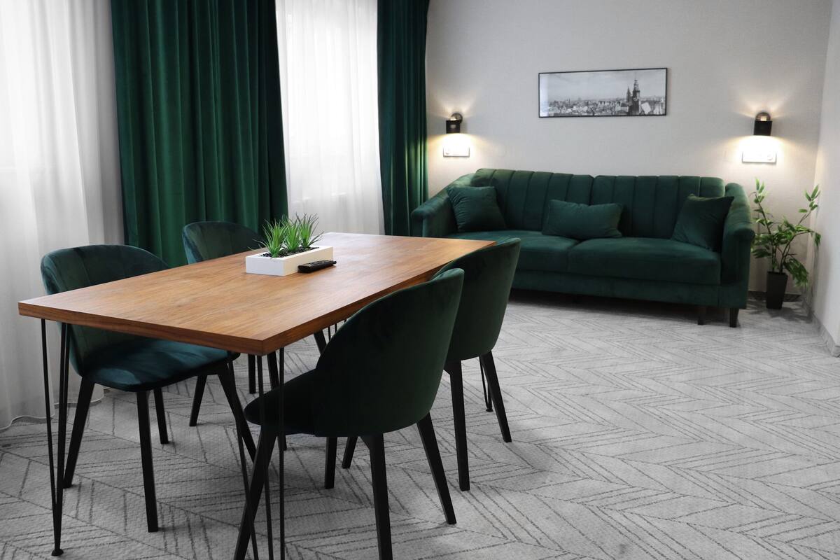 A modern living room with a wooden dining table, green upholstered chairs, a green sofa, and two wall-mounted lights.
