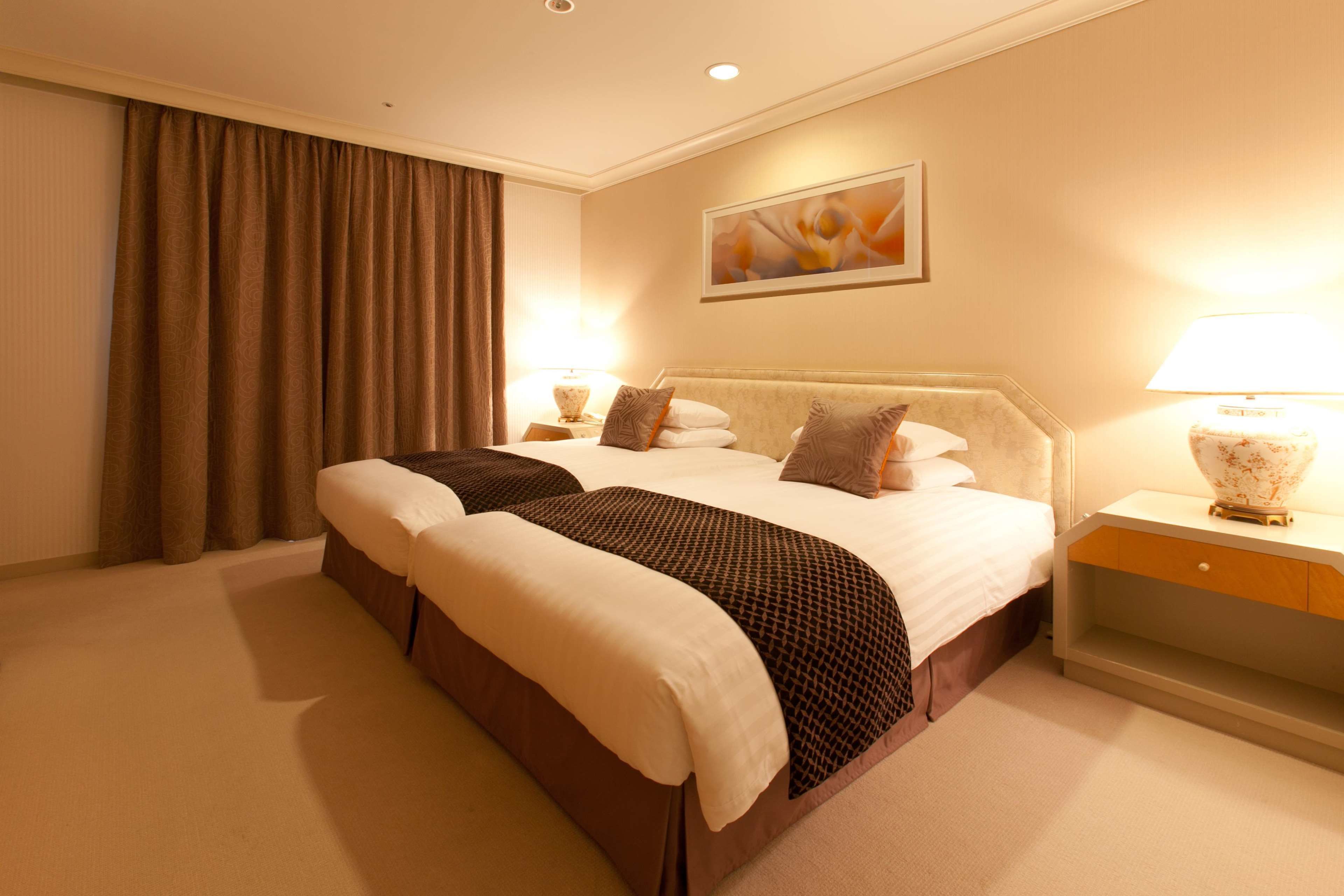 A hotel room with a double bed, two bedside tables, a lamp, a painting on the wall, and a large window with curtains.