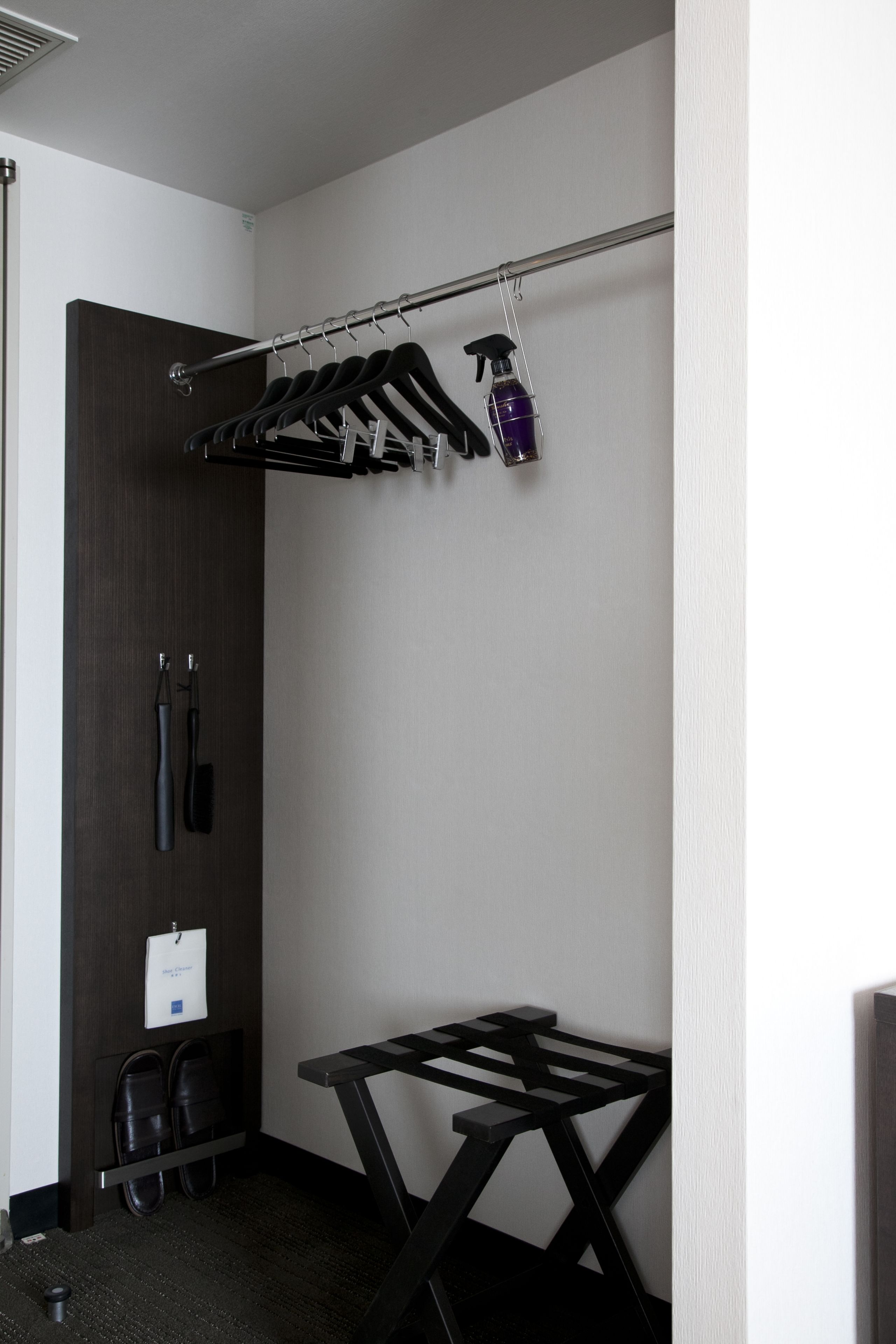 A hotel room with a metal rack holding several black hangers, a spray bottle, and a small whiteboard on the wall. There is a black folding table and a pair of black shoes on the floor.