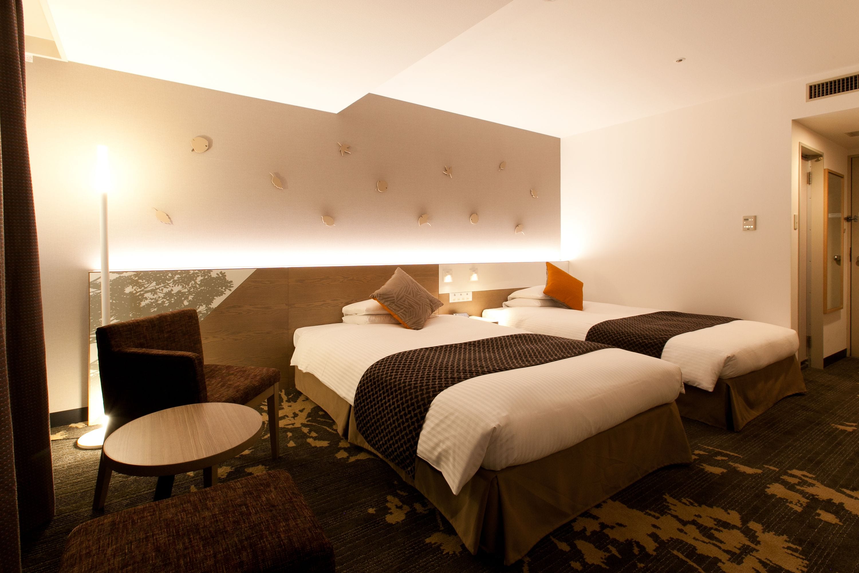 Una habitación de hotel con dos camas, una silla, una mesita y una lámpara fijada a la pared.