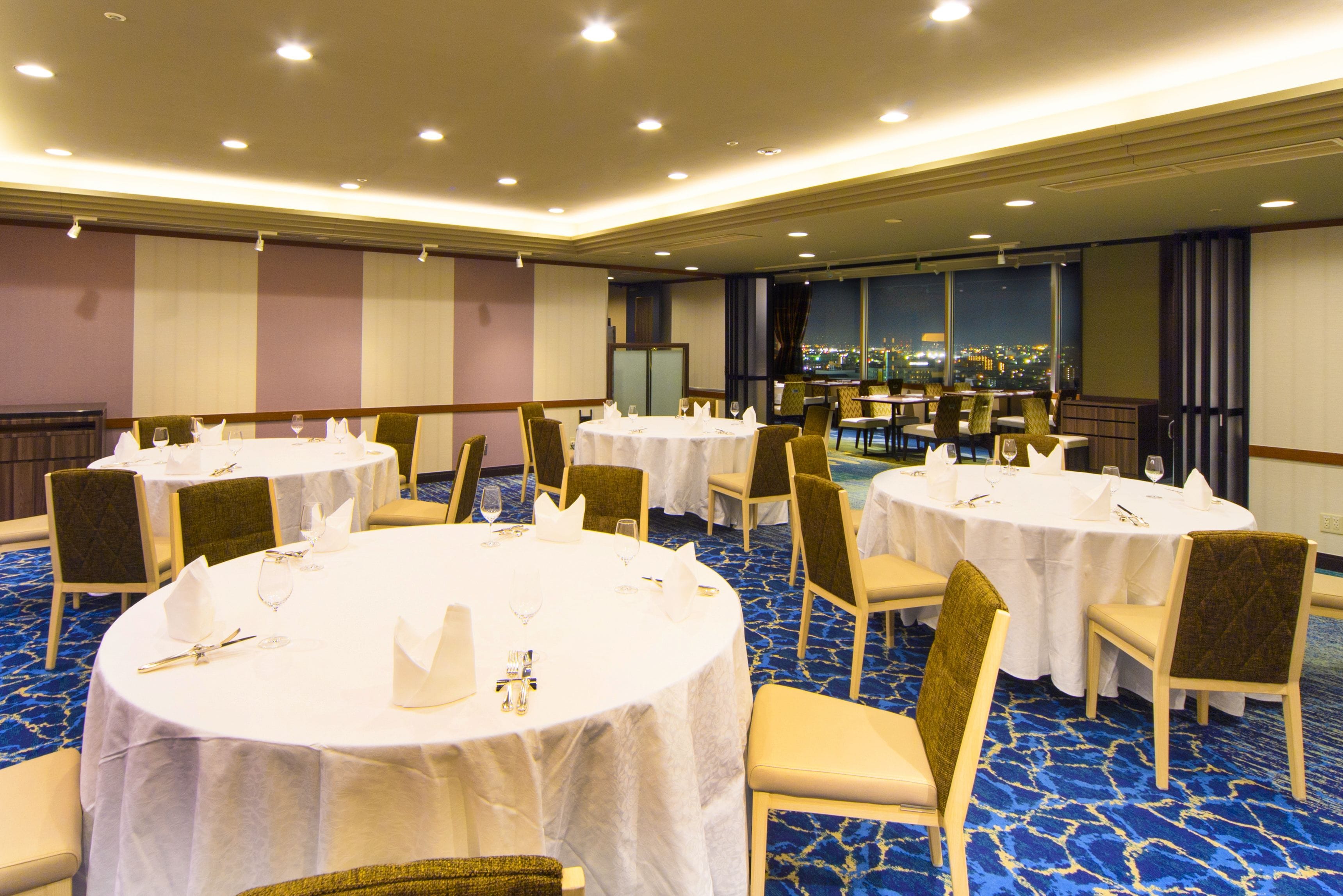 A banquet hall with multiple round tables set for dining, each with white tablecloths and chairs, and a cityscape view through large windows.